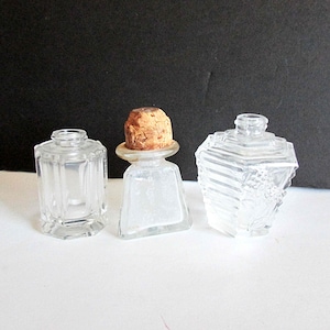May include: Three clear glass bottles with stoppers. The bottle on the left has a square shape with a faceted design. The bottle in the middle has a square base and a square top with a cork stopper. The bottle on the right has a hexagonal shape with a faceted design.