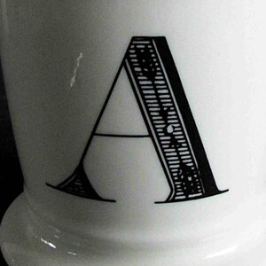 Op de afbeelding: Witte keramische mok met een grote, zwarte letter "A". De letter heeft een gedetailleerd ontwerp aan de rechterkant. De mok heeft een afgeronde basis en een gladde, glanzende afwerking.