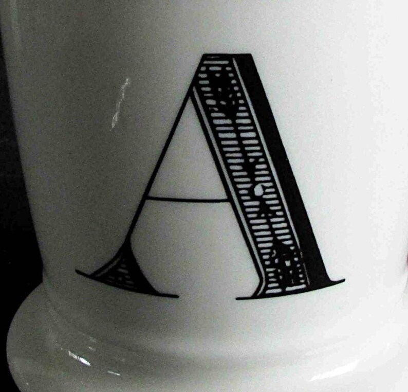 Op de afbeelding: Witte keramische mok met een grote, zwarte letter "A". De letter heeft een gedetailleerd ontwerp aan de rechterkant. De mok heeft een afgeronde basis en een gladde, glanzende afwerking.