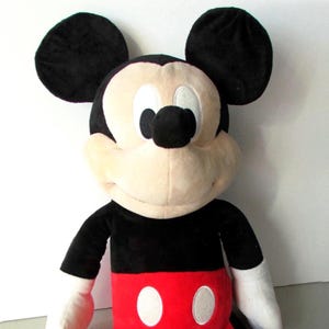 May include: A plush Mickey Mouse toy. The toy features large black ears, a black body, white gloves, and red shorts with white buttons. The face is a light tan color with black eyes and nose. The toy is soft and cuddly.