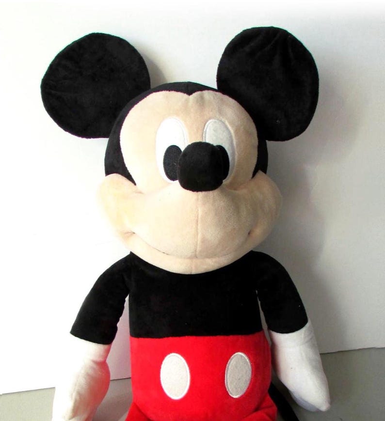 May include: A plush Mickey Mouse toy. The toy features large black ears, a black body, white gloves, and red shorts with white buttons. The face is a light tan color with black eyes and nose. The toy is soft and cuddly.