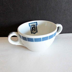 May include: White ceramic cup with two handles and a blue square pattern around the rim. The interior features a blue and black crest design. The cup is set against a white surface and a black background.