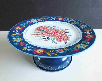 Könnte beinhalten: Ein blauer Kuchenständer aus Metall mit floralem Design. Der Ständer hat einen weißen Teller mit rosa und orangefarbenen Blumen in der Mitte, umgeben von einem blauen Rand mit bunten floralen Akzenten. Der Sockel ist ebenfalls blau.