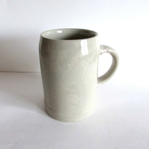May include: A white ceramic beer stein with a handle. The stein has a smooth, slightly textured surface.