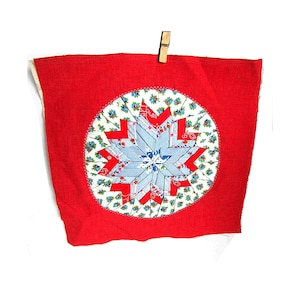 May include: A red fabric square with a central starburst design. The starburst is composed of light blue and red fabric pieces, surrounded by a floral patterned circle. A wooden clothespin holds the fabric.