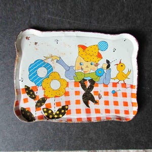 May include: A vintage metal tray with a checkered pattern in orange and white. The tray features a cartoon illustration of a person with a blue shirt and a yellow hat, holding a blue flower. A yellow bird is perched on a branch to the right of the person.