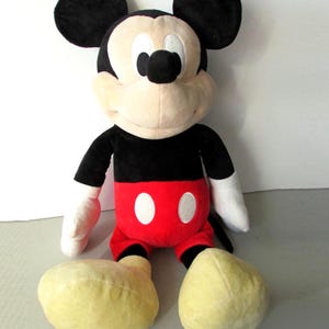 May include: A plush Mickey Mouse toy. The iconic character is depicted with black ears, a black torso, red shorts with white buttons, white gloves, and large yellow feet. The toy is soft and cuddly, perfect for children and Disney fans.