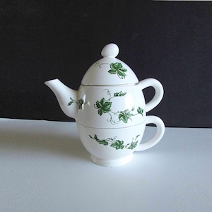 May include: White ceramic tea-for-one set with a teapot and cup design. The set features green leaf and vine patterns, with a lid and handle on the teapot. The cup is stacked beneath the teapot, creating a compact and elegant design.