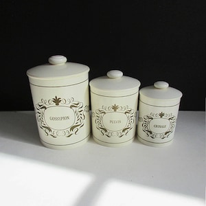 May include: Three off-white ceramic canisters with lids, labeled "GOSSYPION", "PULVIS", and "CRINALE" in a decorative gold font. Each canister has a gold ornate design around the label. The canisters are of varying sizes.