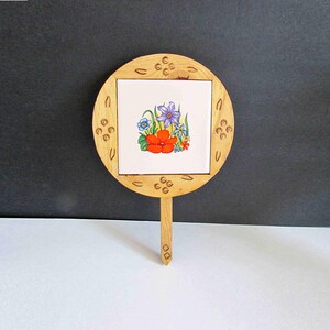 May include: A wooden garden stake with a round frame and a square, floral-patterned tile. The tile features a vibrant arrangement of red, purple, and blue flowers. The stake is made of light-colored wood.