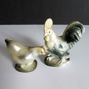 Może przedstawiać: Ceramiczna figurka koguta w kolorze białym z czarnym i zielonym ogonem oraz ceramiczna figurka kury w kolorze białym z szarym łbem. Obie figurki stoją na białym tle.