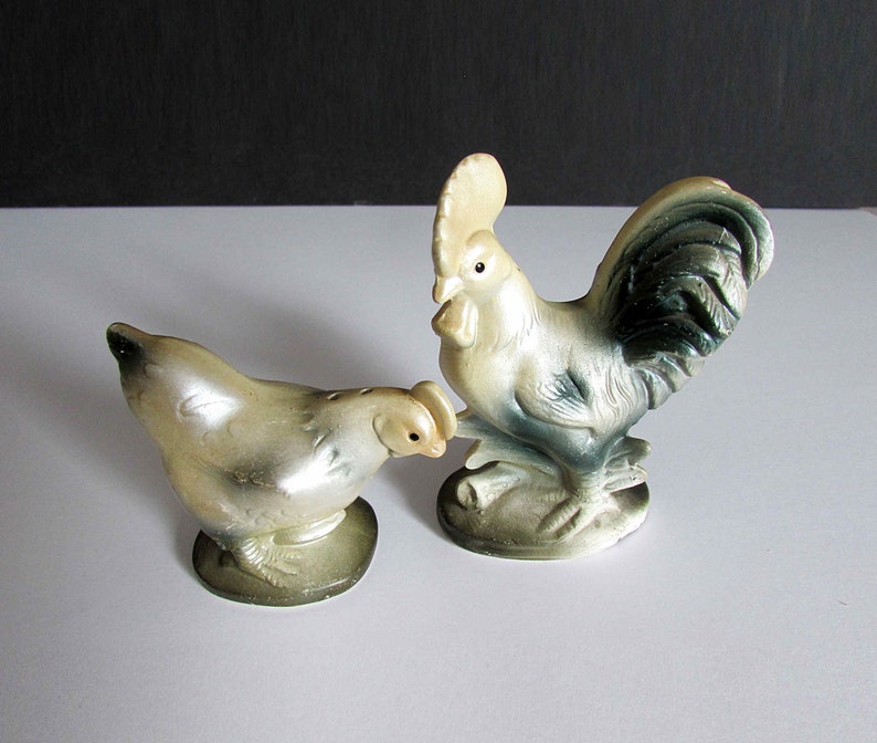 Może przedstawiać: Ceramiczna figurka koguta w kolorze białym z czarnym i zielonym ogonem oraz ceramiczna figurka kury w kolorze białym z szarym łbem. Obie figurki stoją na białym tle.