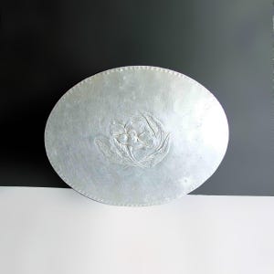 May include: A silver-colored, oval-shaped metal lid with a floral design in the center. The lid has a textured surface and a decorative edge. The floral design appears to be an embossed flower with leaves.