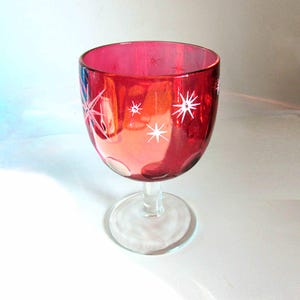May include: A red glass goblet with a clear stem and base. The goblet has a white starburst design.