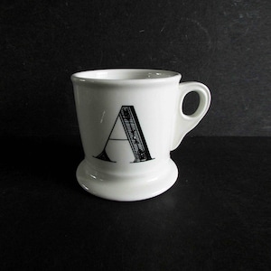 Op de afbeelding: Een witte keramische mok met de zwarte letter "A" erop. De mok heeft een afgeronde basis en een klein handvat. De letter "A" is gestileerd met een decoratief, getextureerd ontwerp.