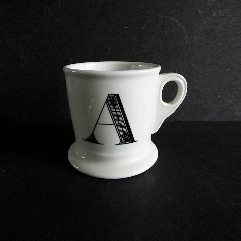 Op de afbeelding: Een witte keramische mok met de zwarte letter "A" erop. De mok heeft een afgeronde basis en een klein handvat. De letter "A" is gestileerd met een decoratief, getextureerd ontwerp.