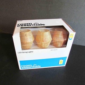 May include: A white cardboard box containing Room Essentials LED string lights. The packaging features a clear plastic window displaying three light-colored, textured, barrel-shaped light covers. The box includes the text Room Essentials and LED String Lights.