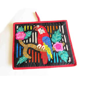 May include: A colorful embroidered wall hanging featuring a parrot perched on a branch with flowers. The parrot is red, yellow, blue, and green. The background is black with colorful stripes. The hanging has a red border.