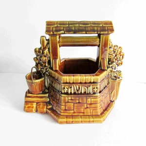 May include: A ceramic wishing well planter in shades of brown and tan. The well features a brick-like design, a small bucket, and decorative floral accents. The roof and the top of the well are a darker brown.