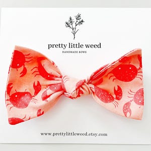 May include: A handmade bow tie with a crab pattern in red and peach. The bow tie is on a white card with the text "pretty little weed" and "handmade bows". The website address www.prettylittleweed.etsy.com is also visible.
