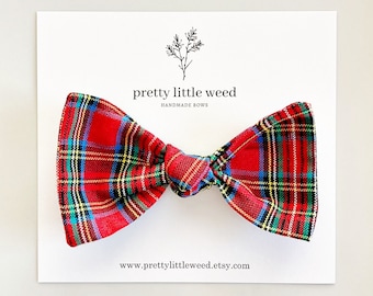 May include: A red plaid bow tie featuring intersecting lines of blue, yellow, and black. The bow is attached to a white display card with the text "pretty little weed HANDMADE BOWS" and "www.prettylittleweed.etsy.com" printed in black ink.