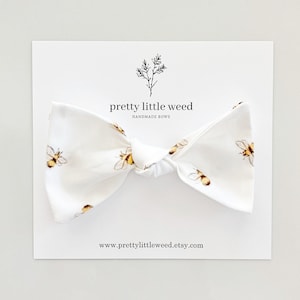 May include: White bow tie with a bee pattern. The bow tie is tied and ready to wear. The bow tie is on a white card with the text "pretty little weed handmade bows" and "www.prettylittleweed.etsy.com".