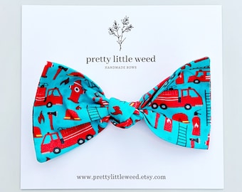 May include: A turquoise fabric bow featuring a repeating pattern of red fire trucks, fire hydrants, ladders, and helmets. The bow is attached to a white display card printed with the text pretty little weed HANDMADE BOWS and www.prettylittleweed.etsy.com.