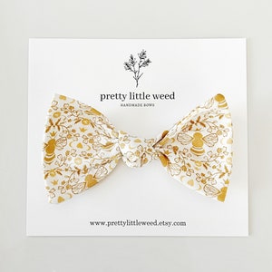May include: A white bow with a gold floral pattern featuring bees, hearts, and moons. The bow is tied and ready to be worn. The text "pretty little weed handmade bows" and "www.prettylittleweed.etsy.com" are printed on the white card behind the bow.