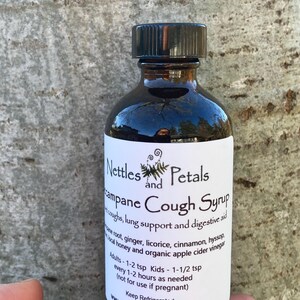 May include: A dark brown glass bottle with a black cap, labeled "Nettles and Petals Campane Cough Syrup". The label lists ingredients like ginger, licorice, and cinnamon, and instructions for use. The bottle is sitting on a tree trunk.