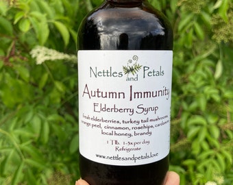 May include: A glass bottle of elderberry syrup labelled "Nettles and Petals Autumn Immunity Elderberry Syrup". The label lists the ingredients: fresh elderberries, turkey tail mushrooms, orange peel, cinnamon, rosehips, cardamom, local honey, and brandy. The label also includes instructions to take 1 tablespoon 1-3 times per day and to refrigerate the syrup. The bottle is held in a hand against a green leafy background.