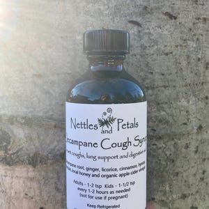 May include: A dark brown glass bottle with a black cap, labeled "Nettles and Petals  Campane Cough Syrup". The label lists ingredients including licorice, cinnamon, and honey. The bottle is held in a hand against a tree bark background.
