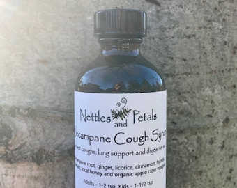 May include: A dark brown glass bottle with a black cap, labelled "Nettles and Petals Campane Cough Syrup". The label lists ingredients including licorice, cinnamon, and honey. The bottle is held in a hand against a tree bark background.
