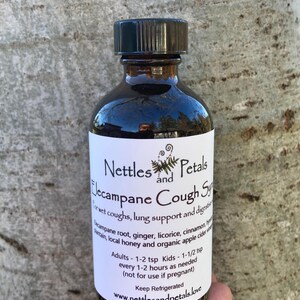 May include: A dark brown glass bottle with a black lid, labeled "Nettles and Petals Lecampane Cough Syrup". The label lists ingredients including elecampane root, ginger, licorice, cinnamon, honey, and apple cider vinegar. It also includes dosage instructions for adults and children.