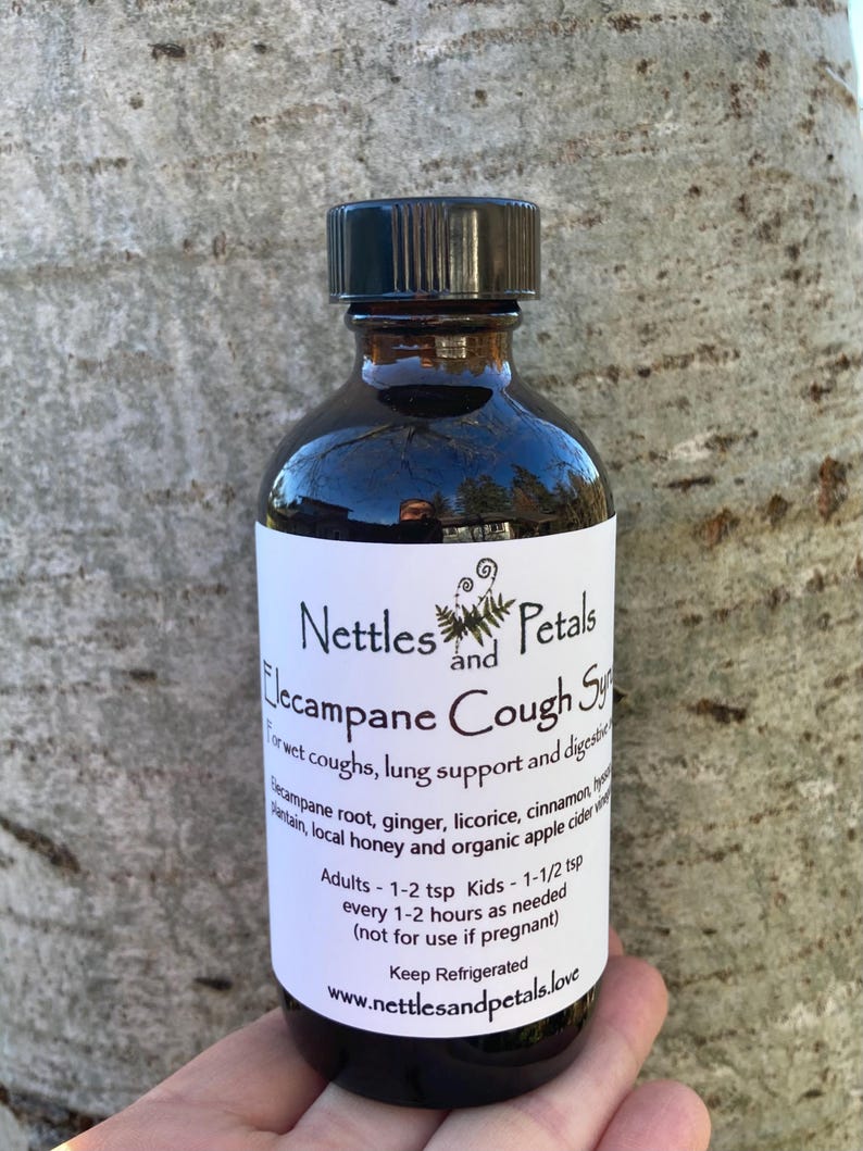 May include: A dark brown glass bottle with a black lid, labeled "Nettles and Petals Lecampane Cough Syrup". The label lists ingredients including elecampane root, ginger, licorice, cinnamon, honey, and apple cider vinegar. It also includes dosage instructions for adults and children.