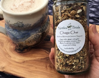 May include: A glass jar of Chaga Chai tea blend with a gold lid, held next to a mug of frothy beverage on a wooden board. The jar's label lists ingredients like chaga, ginger, and cinnamon. The mug has a blue and white wave design.