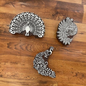 Puede incluir: Tres sellos de bloque de madera con diseños de pavo real. Los sellos son de color marrón oscuro con detalles blancos. Uno es un semicírculo con un pavo real completo, otro tiene forma de lágrima con un diseño estilizado y el tercero es curvo con un pavo real.