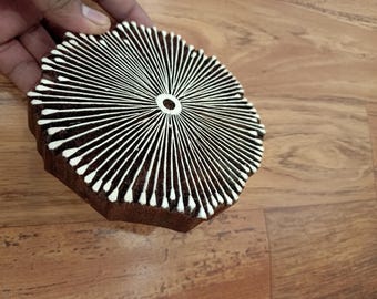 May include: A brown wooden block with a white radial design, held by a hand. The block has a circular center and radiating lines, resembling a sunburst or flower. The block is likely used for printing or stamping.