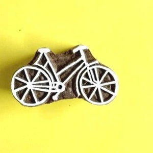 May include: A wooden bicycle stamp with a detailed white design on a brown base. The bicycle features two wheels, a seat, handlebars, and a frame. The stamp is set against a bright yellow background.