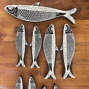 May include: A selection of wooden fish carvings in different sizes, displayed on a wooden surface. The fish are white with black detailing, depicting scales and fins. The carvings vary in size, offering a decorative arrangement.