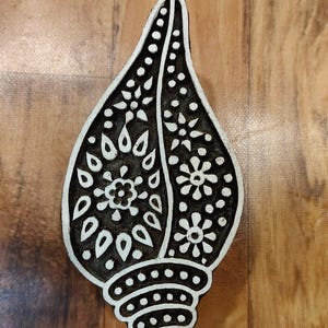 Puede incluir: Un bloque de madera en forma de lágrima en blanco y negro con patrones florales y de puntos. El bloque tiene una base redondeada y probablemente se utiliza para imprimir o estampar diseños en tela o papel. El fondo de vetas de madera proporciona un contraste natural.