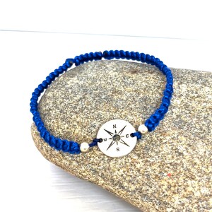 May include: A blue braided bracelet with a silver compass charm. The compass has the directions N, S, E, and W engraved on it.