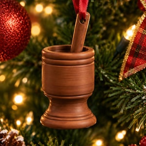 May include: A brown wooden mortar and pestle ornament hanging from a red ribbon. The mortar has a cylindrical body with a flared base and a smooth finish. The pestle is inserted into the top of the mortar, which is suspended against a tree.