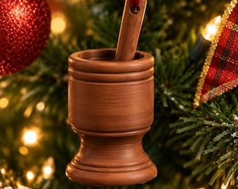 Puede incluir: Un adorno de madera marrón con forma de mortero y mano de mortero colgado de una cinta roja. El mortero tiene un cuerpo cilíndrico con una base ensanchada y un acabado liso. La mano está insertada en la parte superior del mortero.
