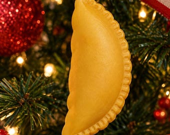 Puede incluir: Un adorno de empanada de color amarillo dorado con un borde rizado, suspendido por un cordón dorado. La réplica de masa en forma de media luna presenta una textura de superficie lisa y está diseñada para colgarse en el árbol.
