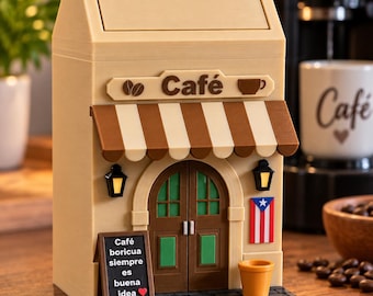 Puede incluir: Un contenedor de basura beige con forma de casa y tapa basculante con la palabra PUSH. El frente tiene un toldo a rayas marrones y blancas, un letrero de Café con iconos de café, una puerta marrón, dos linternas, una bandera de Puerto Rico y un letrero que dice Café boricua siempre es buena idea.