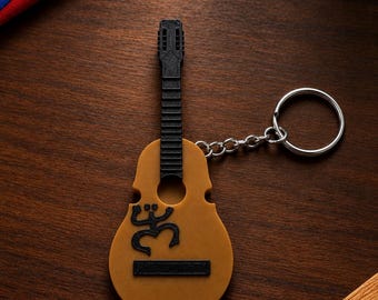 Puede incluir: Un llavero con forma de guitarra que presenta un cuerpo color canela y un mástil negro. El cuerpo muestra un símbolo negro de rana Coquí y un puente rectangular negro. Una anilla de metal plateado está unida a la parte superior con una cadena.