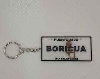 Puede incluir: Un llavero rectangular con fondo blanco y borde negro. El texto dice PUERTO RICO en la parte superior, BORICUA en el centro e ISLA DEL ENCANTO en la parte inferior. Un gráfico marrón de una garita es visible detrás del texto.