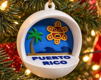 Puede incluir: Un adorno navideño esférico blanco con un diseño recortado en capas de una palmera verde, olas azules y un símbolo de sol amarillo y rojo. El texto PUERTO RICO está impreso en letras azules en la base frontal del adorno.
