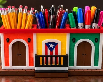 Puede incluir: Un organizador de escritorio rectangular con forma de edificio de tres secciones con paredes naranjas, amarillas y verdes. La sección central presenta una bandera de Puerto Rico y un balcón negro. El organizador está lleno de lápices y rotuladores.