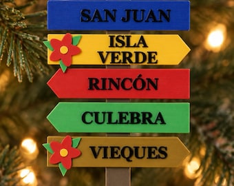 Puede incluir: Un adorno en forma de señal direccional colgado de una cinta roja. El letrero tiene cinco tablones horizontales con los nombres San Juan, Isla Verde, Rincón, Culebra y Vieques en negro, decorado con dos flores rojas.